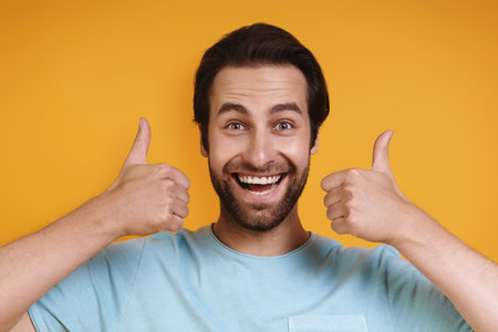 Portrait of happy young man in casual clothing looking at camera and showing thumbs up while standing against yellow backgroundの写真素材