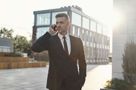 Confident mature businessman talking on mobile phone while walking near office buildingの写真素材