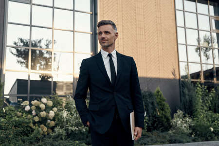 Confident mature man in full suit carrying laptop while standing near office building outdoorsの写真素材
