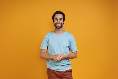 Handsome young man in casual clothing looking at camera and smiling while standing against yellow...