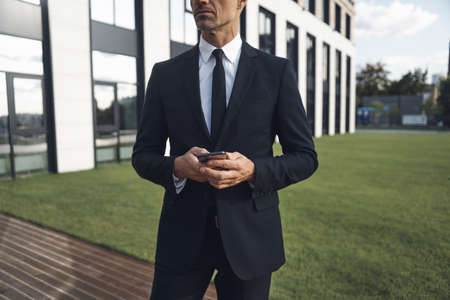Confident mature businessman holding mobile phone while standing near office buildingの写真素材