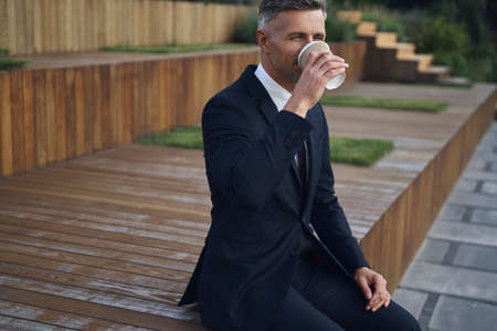 Relaxed mature businessman drinking coffee while sitting outdoors near office buildingの写真素材