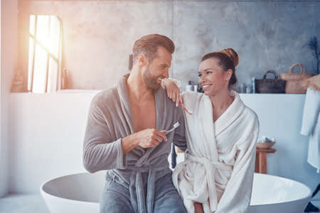 Beautiful young couple in bathrobes smiling and cleaning teeth while doing morning routineの写真素材