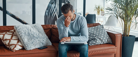 Frustrated young man touching his head and keeping eyes closed while sitting on the sofa at home
