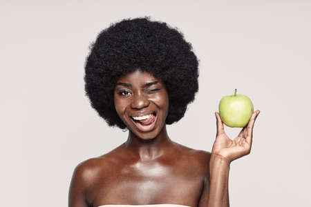 Portrait of beautiful young African woman holding green apple and winkingの写真素材