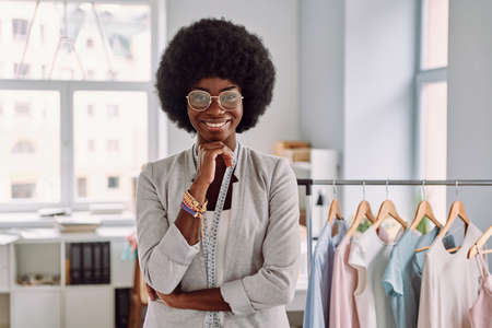 Beautiful young African woman keeping arms crossed and smiling while standing in the fashion design studioの写真素材