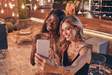 Two beautiful women in evening gowns making selfie and smiling while spending time on luxury partyの写真素材