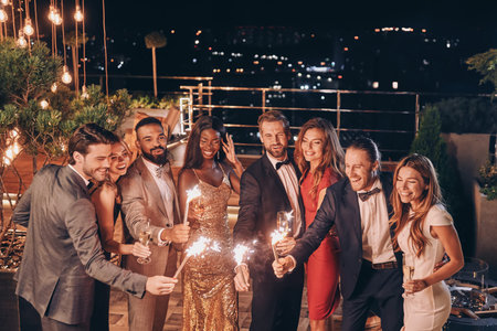 Group of beautiful people in formalwear holding sparklers and smiling while spending time on outdoor...