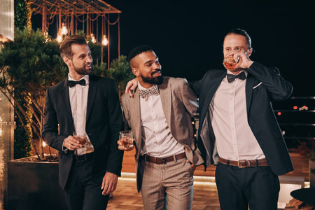 Three handsome men in suits drinking whiskey and communicating while spending time on party