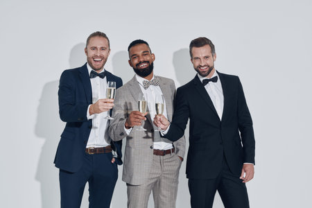 Three handsome young men in suits and bowties toasting with champagne while standing against gray...