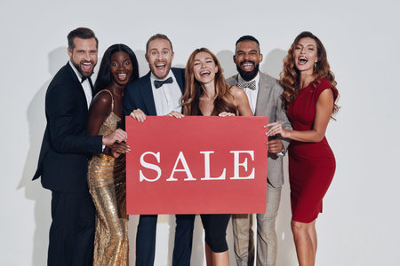 Group of beautiful people in formalwear holding sale banner and smiling while standing against gray...