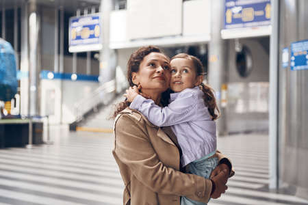 Beautiful mature woman carrying her little daughter and looking away while standing at airport terminalの写真素材