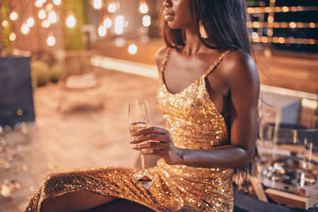 Beautiful young African woman in evening gown holding flute with champagne while spending time on night partyの写真素材