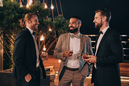 Three handsome men in suits drinking whiskey and communicating while spending time on partyの写真素材