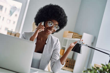 Beautiful young African woman talking on mobile phone and smiling while sitting at her working place in officeの写真素材