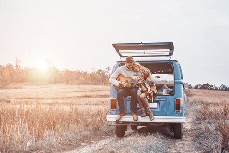 Handsome young man playing guitar for his girlfriend while both sitting in the car trunk outdoors