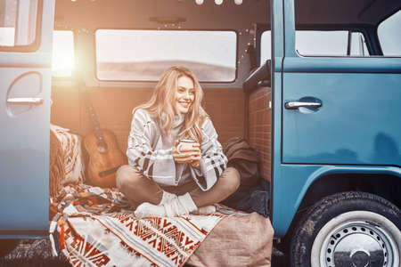 Beautiful young woman enjoying hot drink and smiling while spending time in retro minivanの写真素材