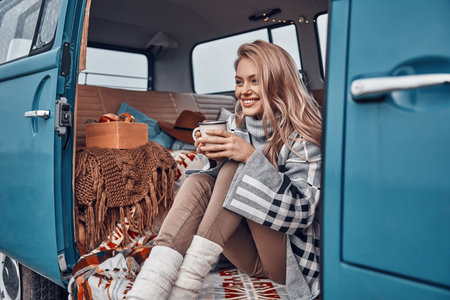 Beautiful young woman enjoying hot drink and smiling while spending time in retro minivan