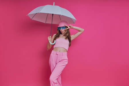 Playful young woman in trendy clothing holding umbrella while standing against pink backgroundの写真素材