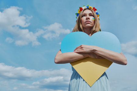 Beautiful Ukrainian woman holding heart shape national flag upon head with sky in the backgroundの写真素材