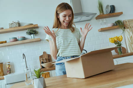 Excited young woman unpacking box while standing at the domestic kitchenの写真素材