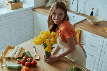 Top view of beautiful young woman holding yellow tulips and smiling while standing at the kitchen counterの写真素材