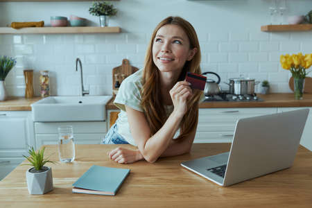 Attractive young woman enjoying online shopping and using credit card while sitting at the kitchenの写真素材