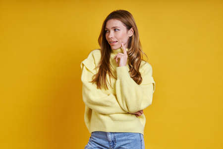 Beautiful young woman holding hand on chin and smiling while standing against yellow backgroundの写真素材