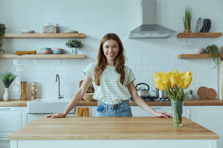 Beautiful young woman leaning at the kitchen desk and smilingの写真素材
