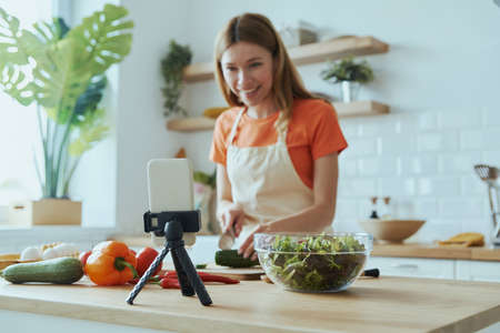Beautiful young woman cooking and vlogging while standing at the domestic kitchenの写真素材