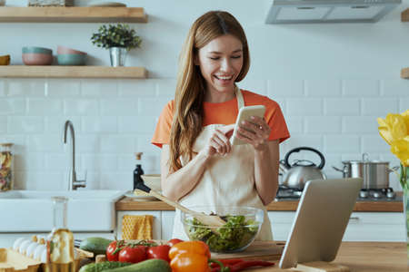 Happy young woman using technologies while cooking at the domestic kitchenの写真素材