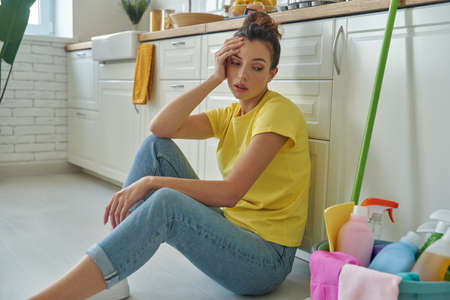 Tired young woman sitting near the bucket with cleaning stuff at the domestic kitchenの写真素材