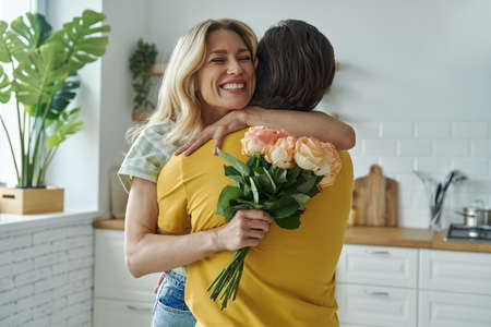Young man surprises his girlfriend with bunch of flowers at the kitchenの写真素材