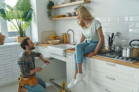 Cheerful young man repairing a sink while woman sitting on the kitchen counterの写真素材