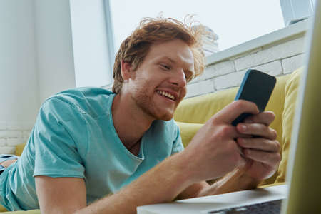 Handsome redhead man using technologies and smiling while lying on the couch at homeの写真素材