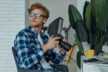 Confident young man holding digital camera while sitting at his working place in studioの写真素材