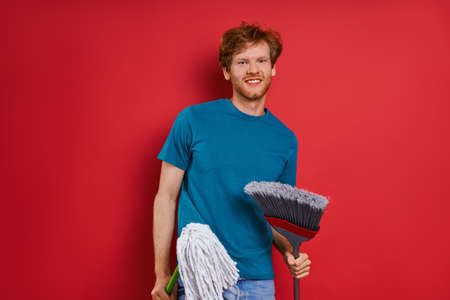 Cheerful young man carrying cleaning equipment while against red backgroundの写真素材