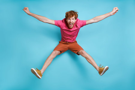 Cheerful young redhead man jumping against blue background