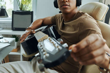 Concentrated young mixed race man tuning acoustic guitar while sitting at his working placeの写真素材