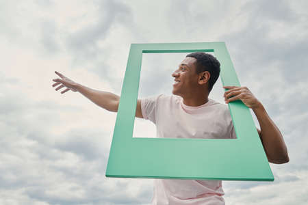 Cheerful mixed race man looking through a picture frame and pointing skyの写真素材