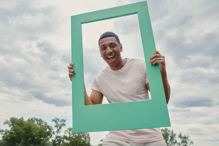 Happy mixed race man looking through a picture frame while standing outdoorsの写真素材