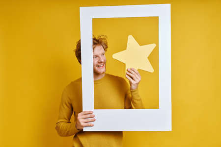 Happy man holding star while looking through a picture frame against yellow backgroundの写真素材