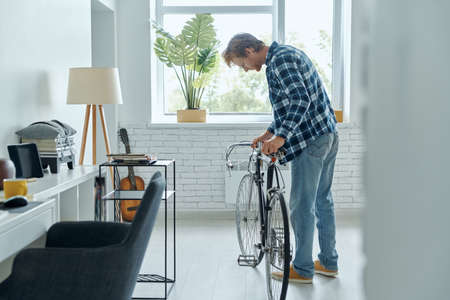 Cheerful young man examining his bicycle while standing at the living roomの写真素材
