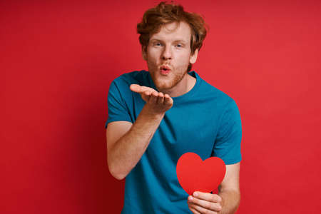 Handsome redhead man holding red paper and blowing a kiss while standing against red backgroundの写真素材