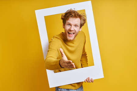Happy man looking through a picture frame and gesturing while standing against yellow backgroundの写真素材