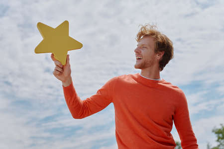 Cheerful young redhead man holding yellow star and smiling with sky in the backgroundの写真素材