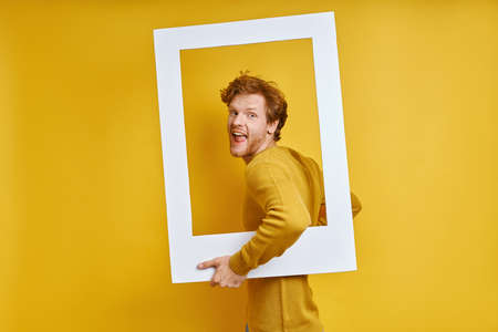 Cheerful redhead man looking through a picture frame while standing against yellow backgroundの写真素材