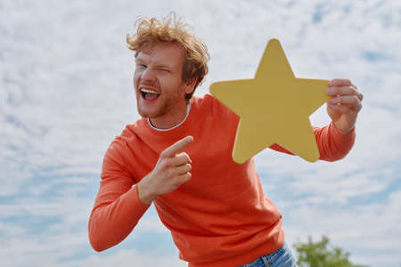 Cheerful young redhead man showing yellow star and smiling with sky in the backgroundの写真素材