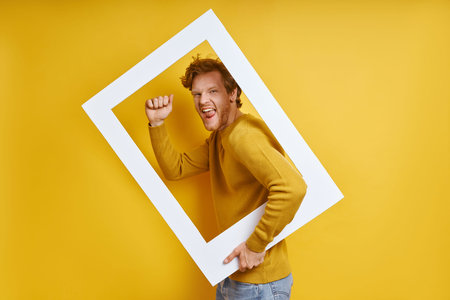 Happy man looking through a picture frame and gesturing while standing against yellow backgroundの写真素材