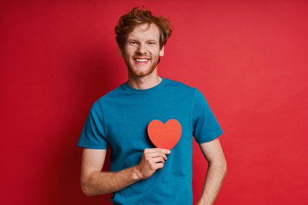 Cheerful redhead man holding red paper heart while standing against red backgroundの写真素材